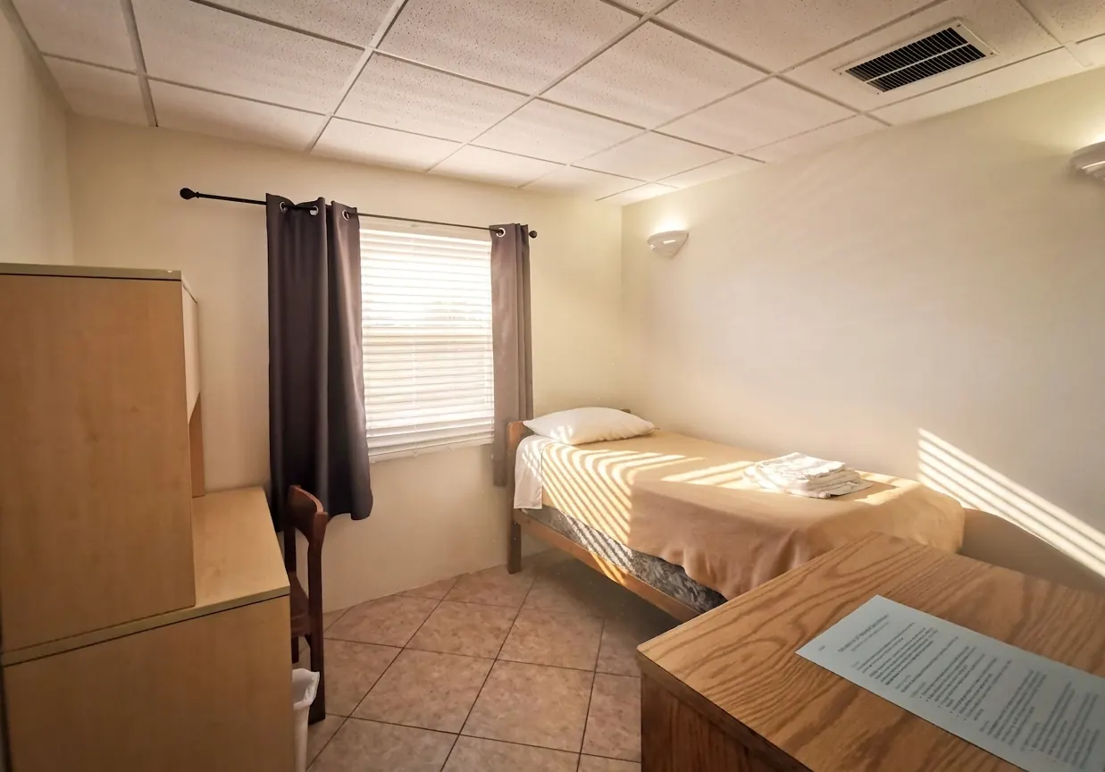 A bedroom with a made bed, a curtained window and a desk along the wall
